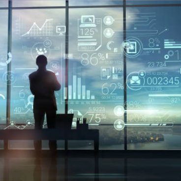 A silhouette of a man stands on the background of large office windows and views a hologram of corporate infographic with work data.
